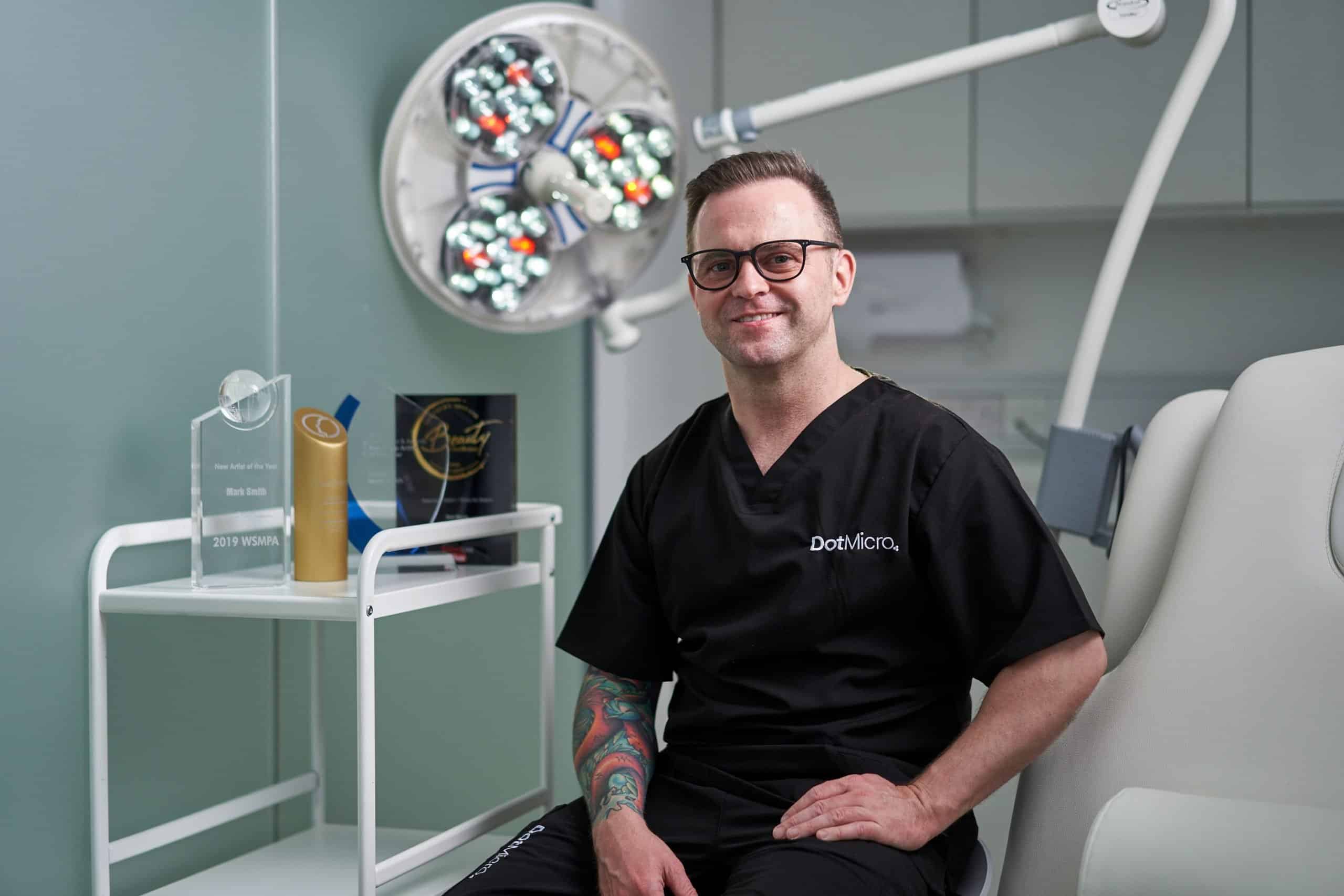 Scalp micropigmentation practitioner seated in a clinical treatment room with equipment visible